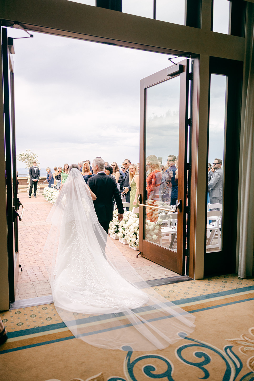 A bride and her father stepping through open doors toward the ceremony as guests stand watching